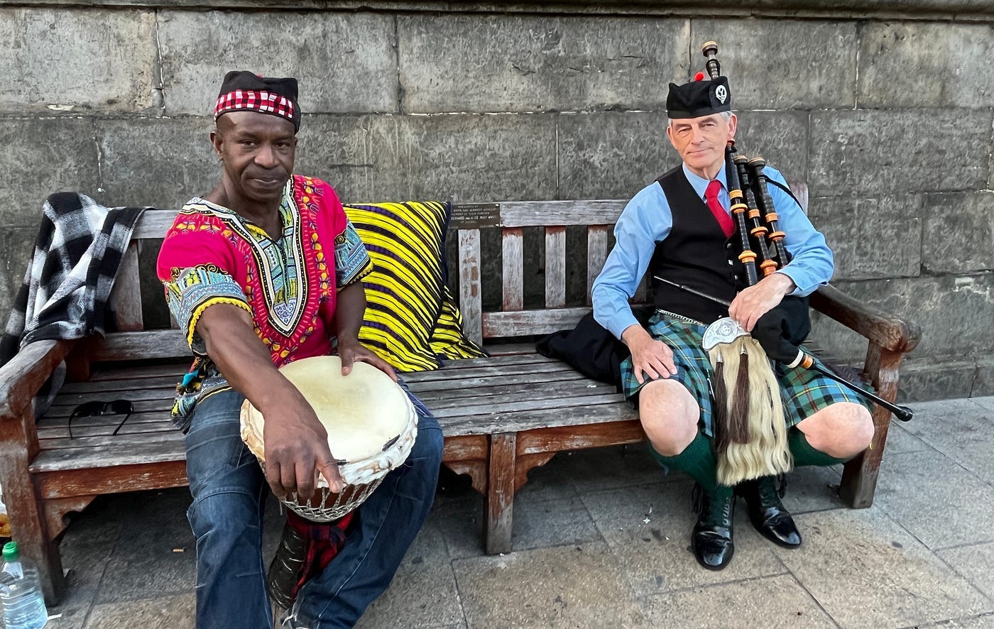 2 gentlemen sit on a bench in front of an old granite wall. On the left, a Black man in a bright, African shirt holding a djembe drum. On the right, a White man in a kilt and Scots regalia holding bagpipes.