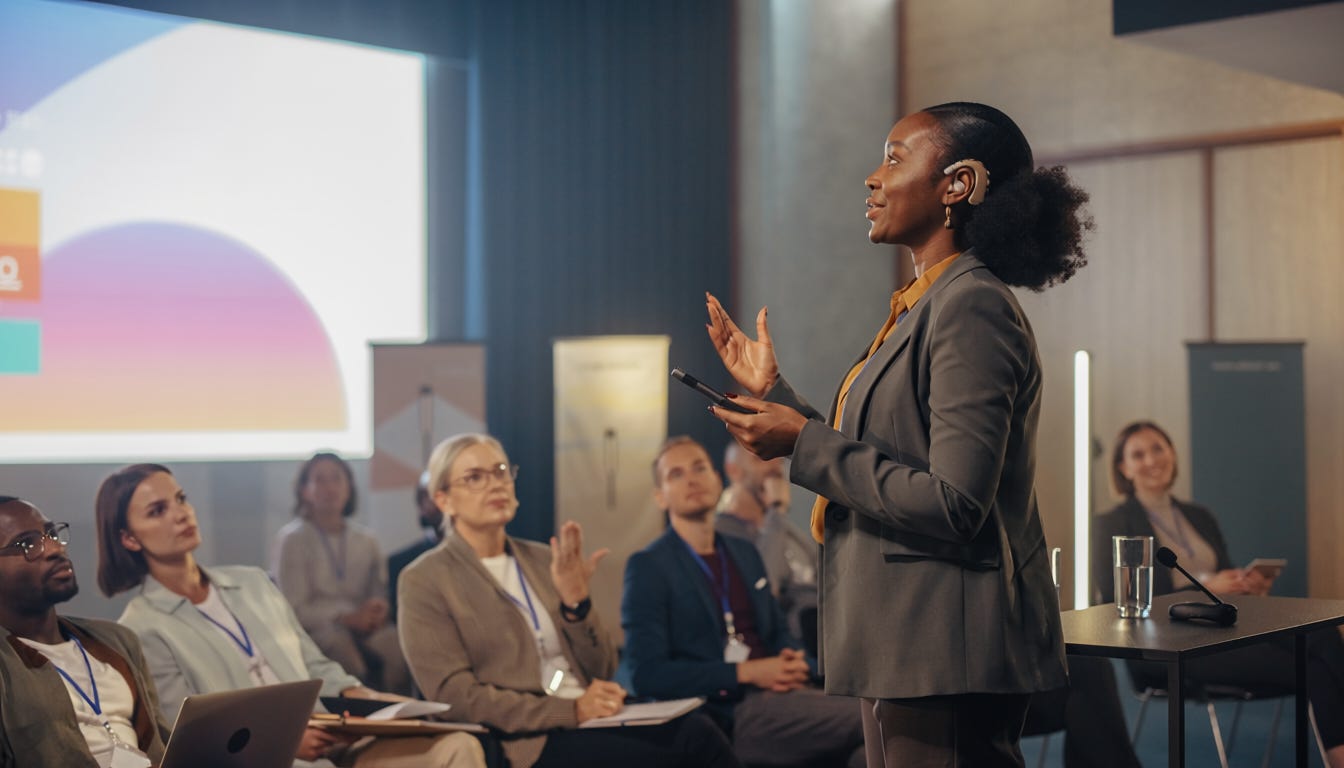 A Black woman wearing a hearing aid presents at a design conference to an engaged audience. A Black woman wearing a hearing aid presents at a design conference to an engaged audience.