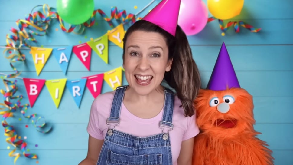 Ms. Rachel and her oragne puppet friend Herbie celebrate a birthday. Ms. Rachel is wearing a pink party hat.