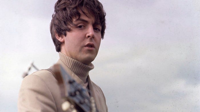A young Paul McCartney, wearing a roll-neck sweater, stands outside playing a guitar