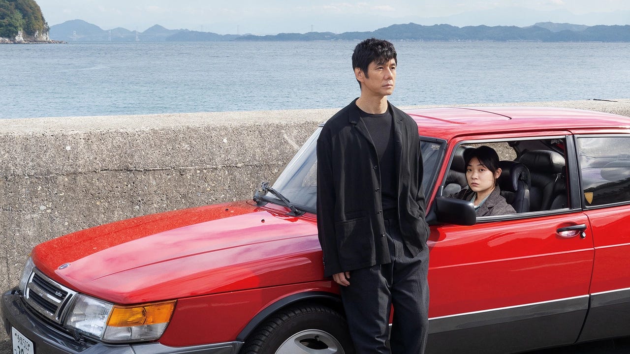 Carro vermelho antigo com uma mulher japonesa ao volante olhando para a câmera. Em frente ao carro tem um homem japonês olhando para o sentido oposto. Ao fundo, se vê o mar.
