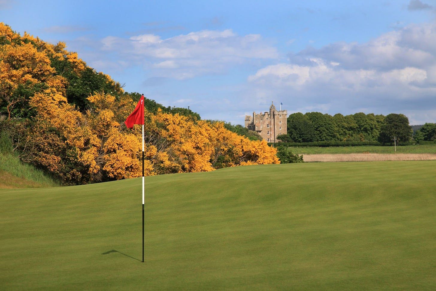 A golf green with a red flag in the foreground, bordered by golden gorse bushes, with a medieval castle visible in the background. A golf green with a red flag in the foreground, bordered by golden gorse bushes, with a medieval castle visible in the background.