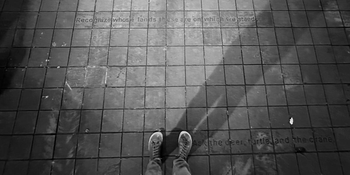 Person stands on sidewalk in a pair of lace-up sneakers. Imprinted in the concrete are the words, "Recognize whose lands these are on which we stand... Ask the deer, turtle, and the crane." Person stands on sidewalk in a pair of lace-up sneakers. Imprinted in the concrete are the words, "Recognize whose lands these are on which we stand... Ask the deer, turtle, and the crane."