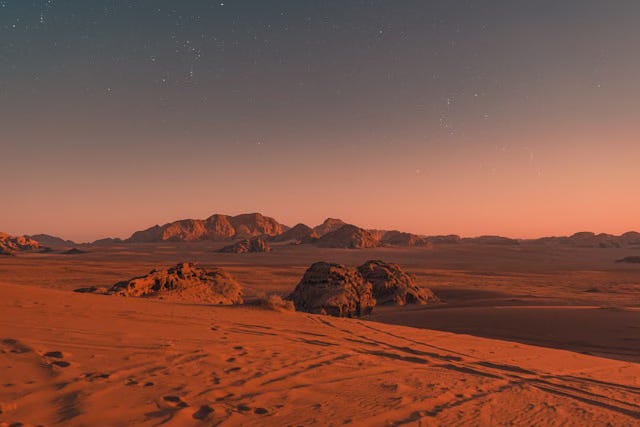 A dark red sandy landscape of Mars with some rover tracks crossing the foreground and low rocky ranges in the distance.