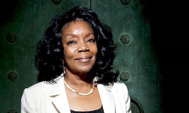 Peggy smiles directly at the camera. She has shoulder length wavy black hair and wears a cream color jacket and a braided pearl necklace. Behind her is a dark teal textured wall.