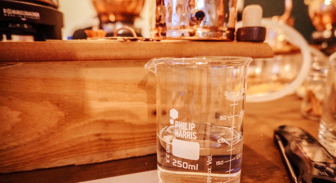 A glass jug with clear rum in it, with a wooden board with a copper distil above it A glass jug with clear rum in it, with a wooden board with a copper distil above it