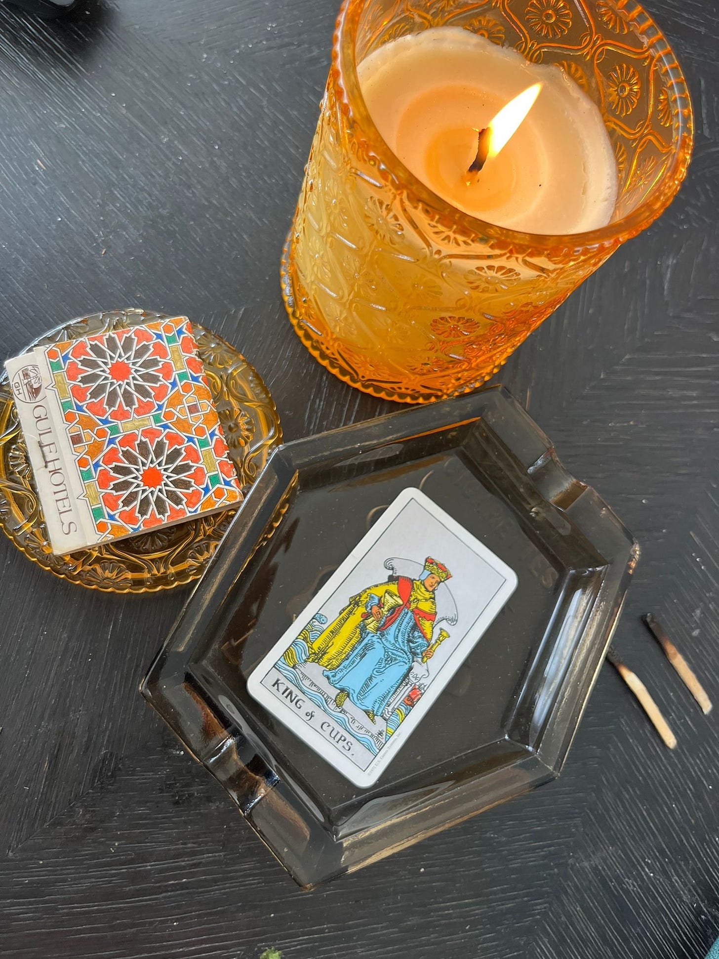The King of Cups sits in an unusual angular ashtray. Two matches lay beside it. A candle is close by and a matchbook rests beside the ashtray, with a mosaic pattern