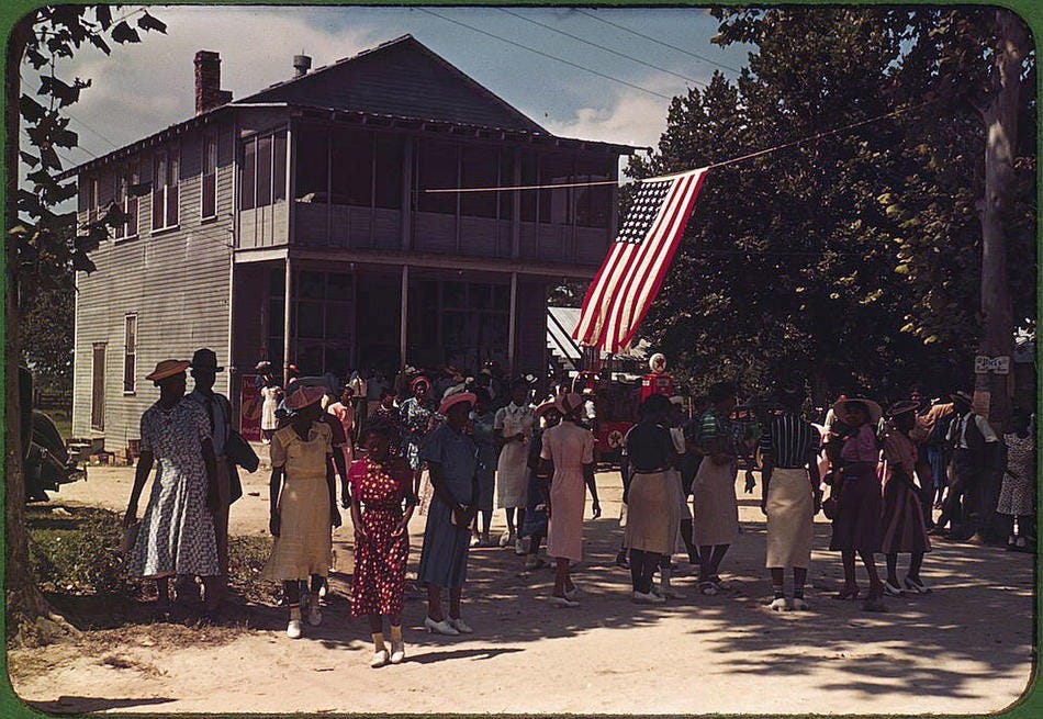 Color photograph of a Fourth of July gathering on a sandy road in front of a two-story white clapboard building with a screened upper porch. A crowd of Black men, women, and children in summer dress fills the road; a woman in a red-and-white polka-dot dress stands at center foreground. A large American flag hangs from a cable strung out from the building, with a filling station visible behind it. Green trees and blue sky fill the background. Color photograph of a Fourth of July gathering on a sandy road in front of a two-story white clapboard building with a screened upper porch. A crowd of Black men, women, and children in summer dress fills the road; a woman in a red-and-white polka-dot dress stands at center foreground. A large American flag hangs from a cable strung out from the building, with a filling station visible behind it. Green trees and blue sky fill the background.