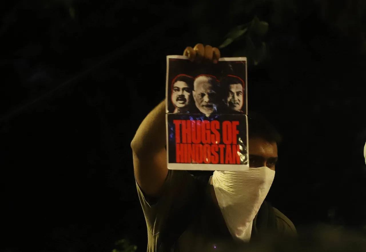 Un manifestante con el rostro cubierto alza una pancarta que muestra a Modi, flanqueado por los ministros Dharmendra Pradhan y Nitin Gadkari, sobre la frase « Los matones de Hindustán » en grandes letras rojas, contra un fondo oscuro.