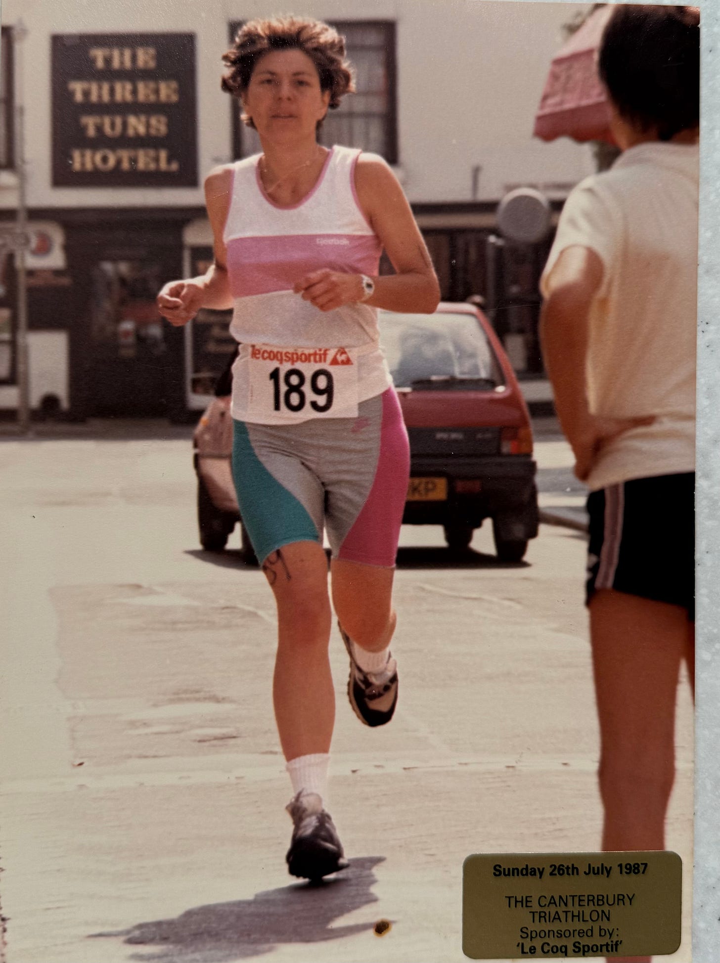 Anne Gould at the end of her first triathlon in 1987. She is wearing a pink and white running vest and grey tri shorts with pink and turquoise panels, soaked and mid-stride, with wet hair and a determined expression after completing the 10K run.