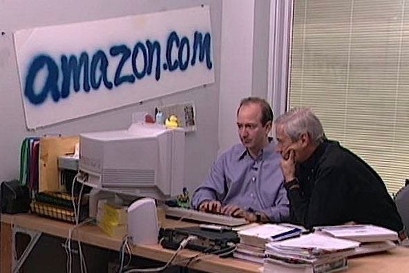 This may contain: two men sitting at a desk in front of an amazon com logo on the wall