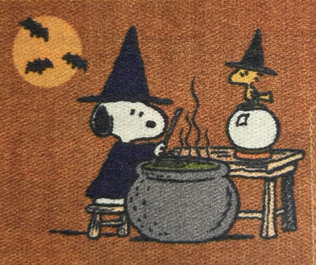 Snoopy wearing a black witch hat, stirring a cauldron with green liquid on a table. Woodstock, also wearing a black witch hat, stands beside a crystal ball on the same table. A full moon with bats is visible in the background.