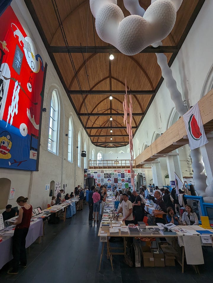 The first image is a poster for the Ghent Art Book Fair, held at Kunsthal Gent on May 11-12, 2024. The poster features bold black text with abstract design elements and lists various participating exhibitors and collaborators, such as Athena! Clay Fair, Imposed Alphabet, and Kero Zine. The second image shows an indoor art book fair scene with high ceilings and colorful tapestries on the walls, featuring multiple booths and tables where people browse through a variety of art books. The space has a grand, church-like architectural style with large windows, wooden beams, and hanging sculptures. In the third image, a person stands behind a booth at the Ghent Art Book Fair, displaying several art books and sticker packs, with a sign indicating that a free sticker pack comes with every book. The table includes works from Soi Books, with a variety of zines, books, and promotional items laid out. The fourth image captures two booth operators from the publishing house Grafis Nusantara sitting behind a table that displays various colorful books, zines, and art publications. One of them is making a peace sign while the other is smiling casually, creating a laid-back and friendly atmosphere at the event.