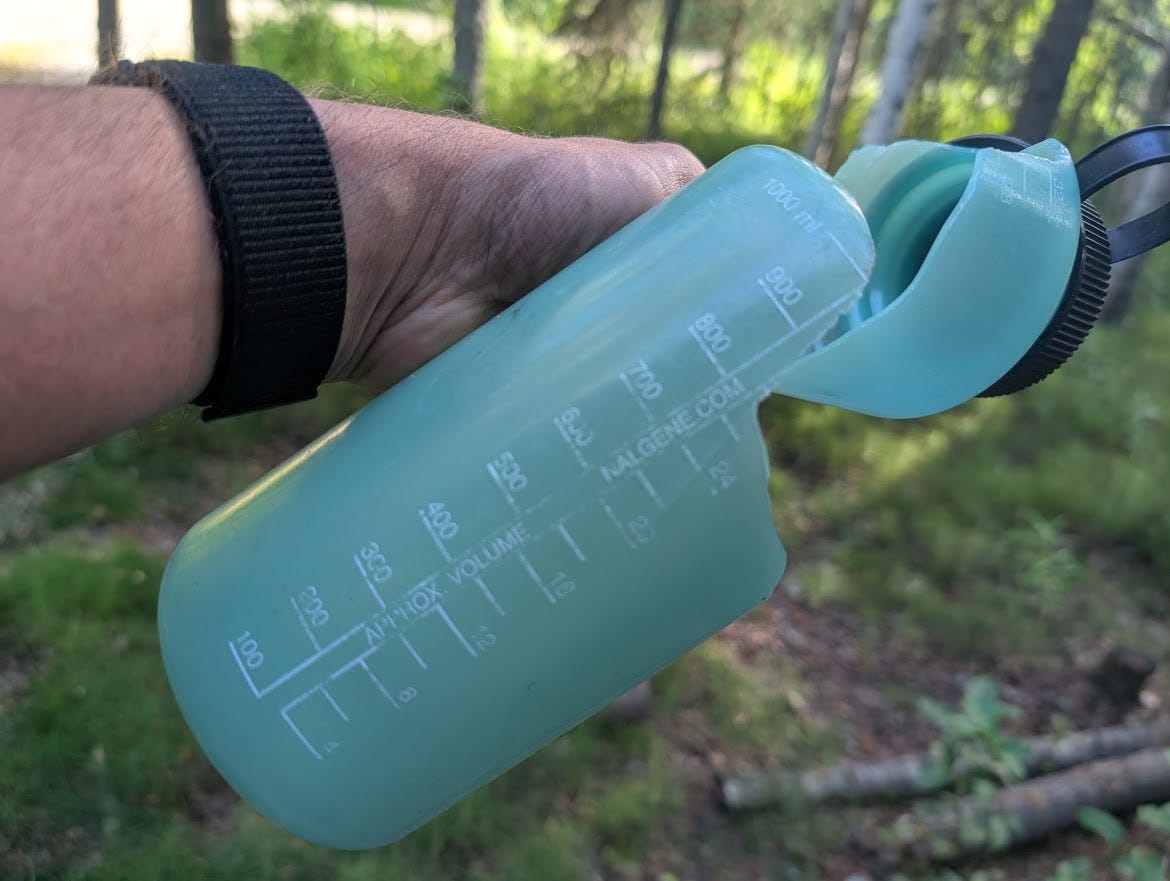 A hand holds up the bottom half of a mint-green Nalgene bottle against a background of sunlit birch forest. The bottle has failed catastrophically: its upper portion is torn and folded over on itself, the black screw cap still attached to the collapsed top, the printed volume markings warped where the plastic deformed. A black watch band circles the wrist holding it.