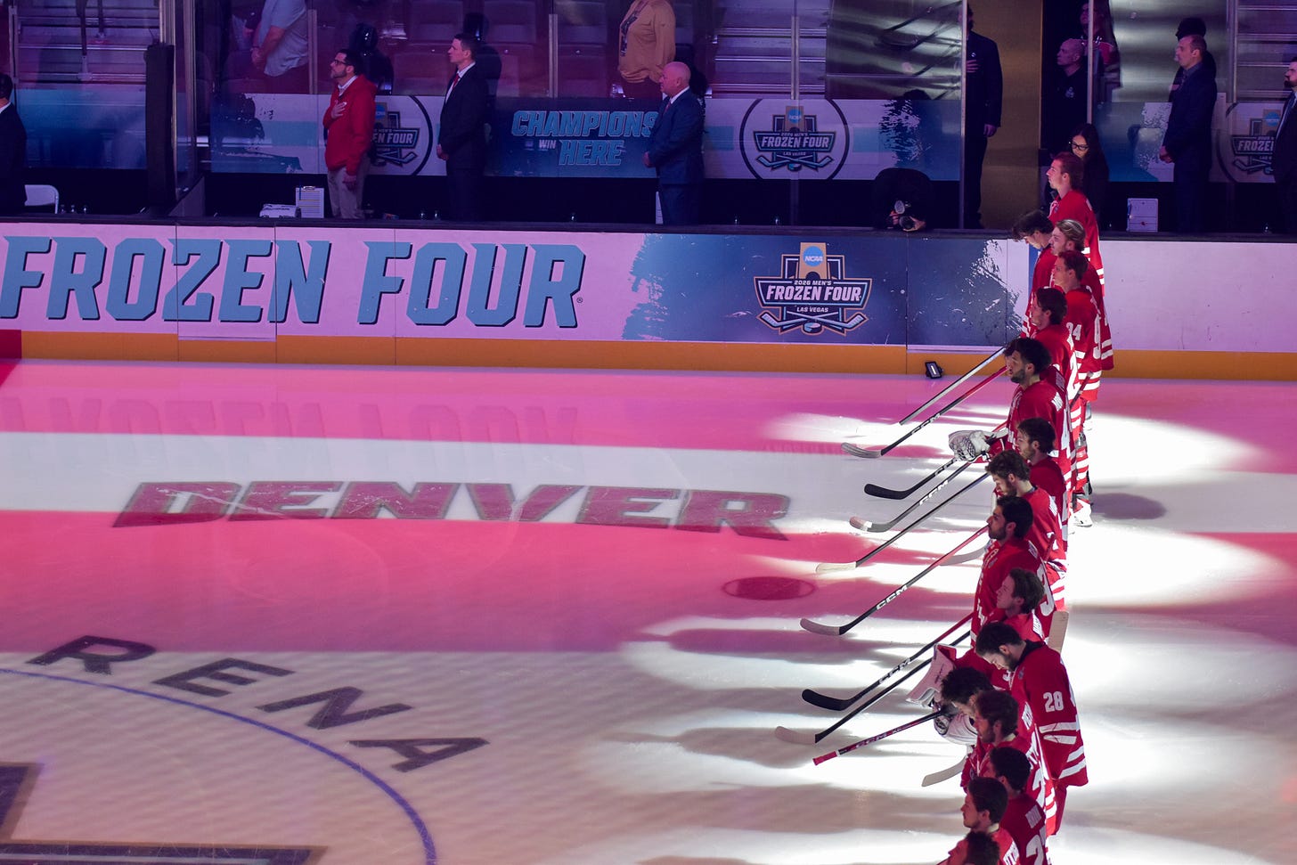 Wisconsin hockey players lined up along blue line under spotlights with 2026 frozen four signage in front of them.
