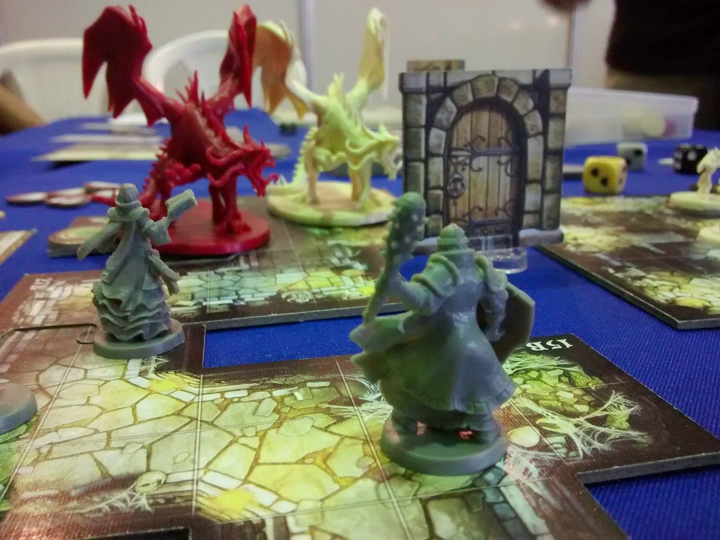 Jogo de tabuleiro com bonecos de plastico cinza, frente uma porta e dois dragoes, um vermelho e um amarelo. Mais ao fundo alguns dados coloridos