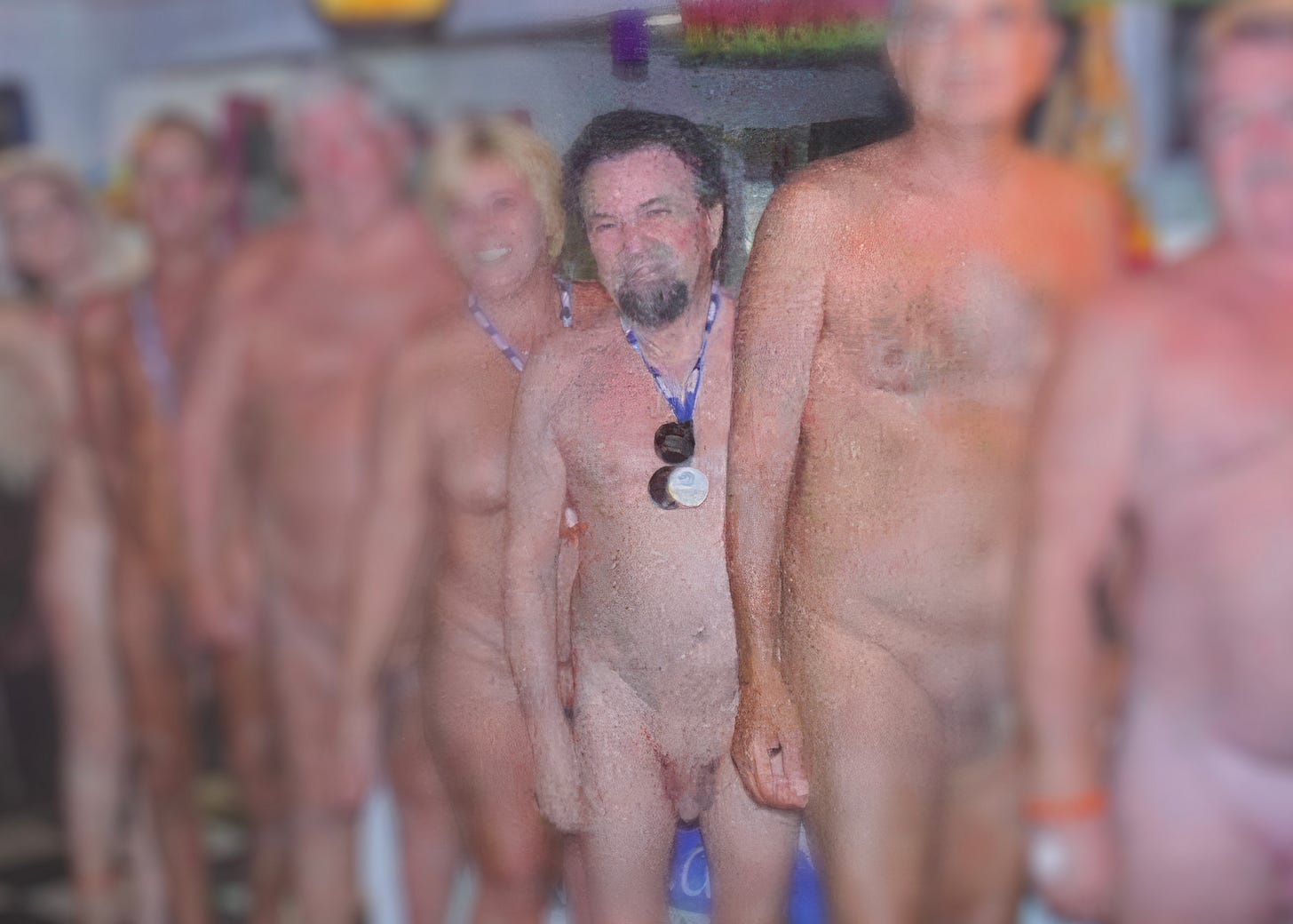 Color photo of a group of nude adults standing in a line at an outdoor or event setting, with one bearded man wearing a medal at center while others stand close beside him, suggesting a group of organizers or officers posing together.