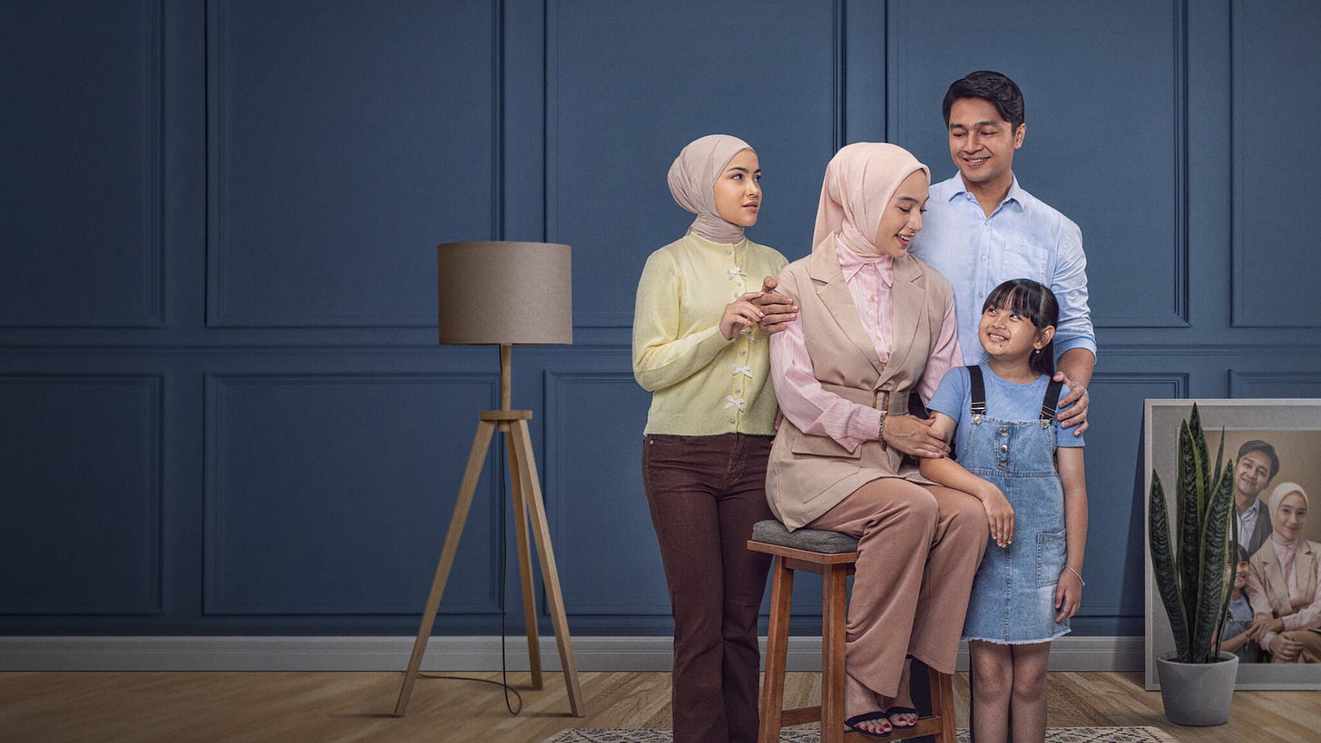 Studio portrait of a Muslim family in a blue-walled living room: two hijab-wearing women stand beside a seated woman holding a young girl, while a man stands behind them smiling. The group appears calm and affectionate, dressed in soft, neutral colors, with a floor lamp and a framed family photo visible in the background.