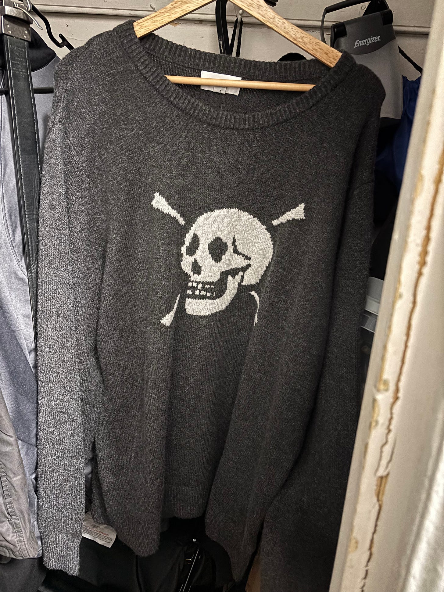 A dark gray knit sweater hanging on a wooden hanger, featuring a white skull and crossbones design on the front.