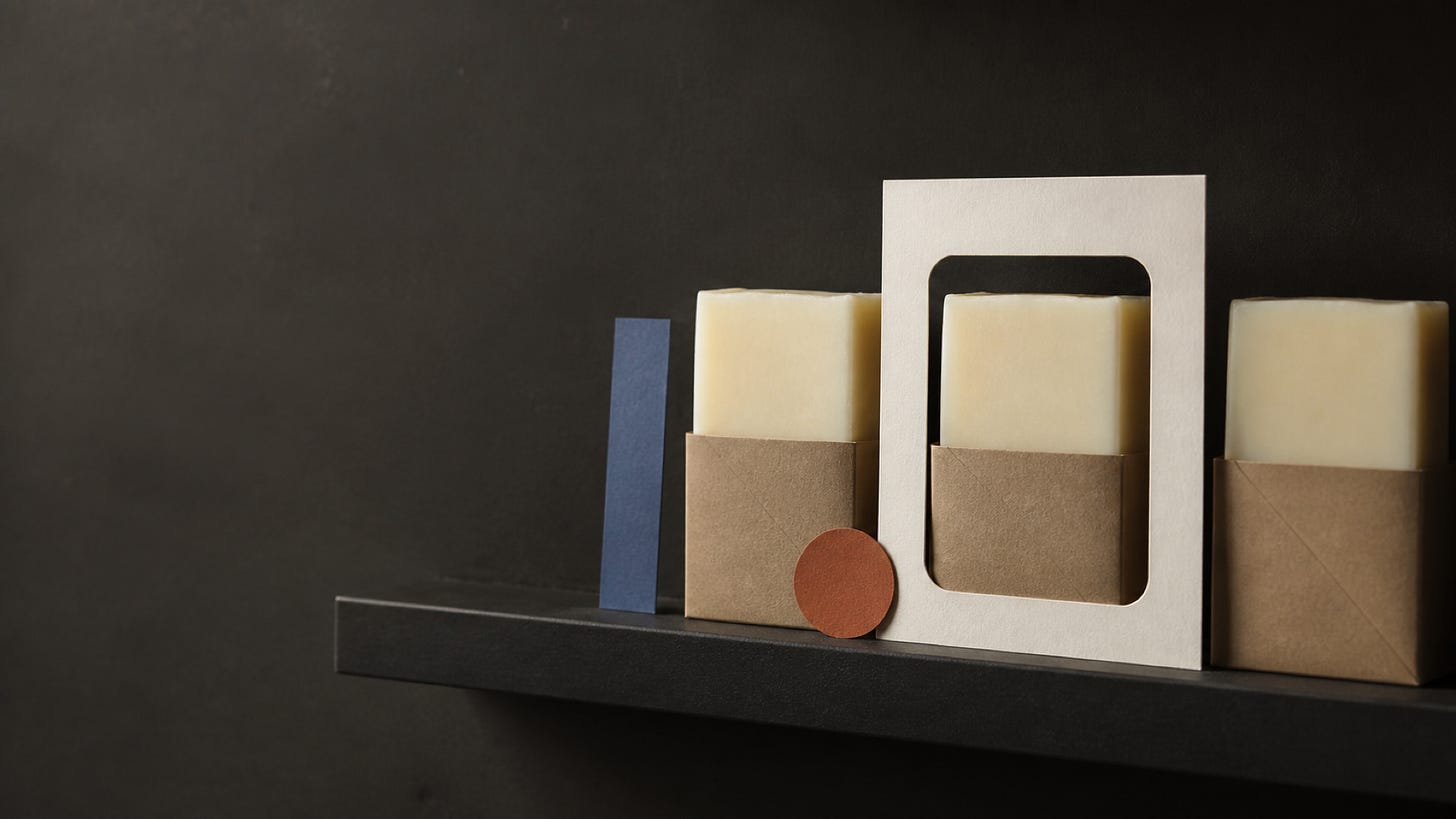 Three rectangular soaps in brown paper wrappers sit on a shelf with geometric paper shapes beside them.