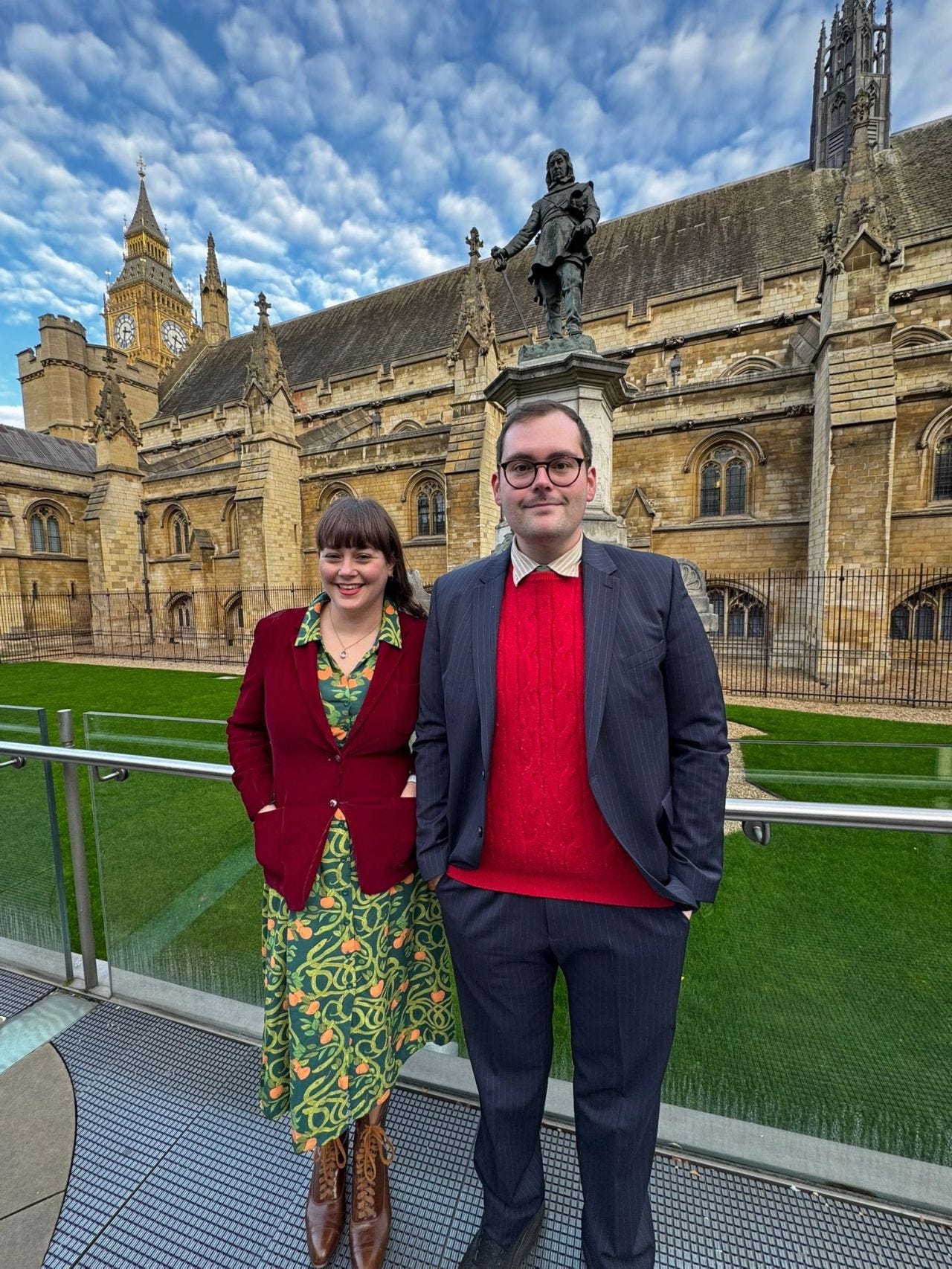 One white woman and one white man stand in smart clothes in front of yellow-bricked Houses of Parliament, with a blue sky with clouds behind. 