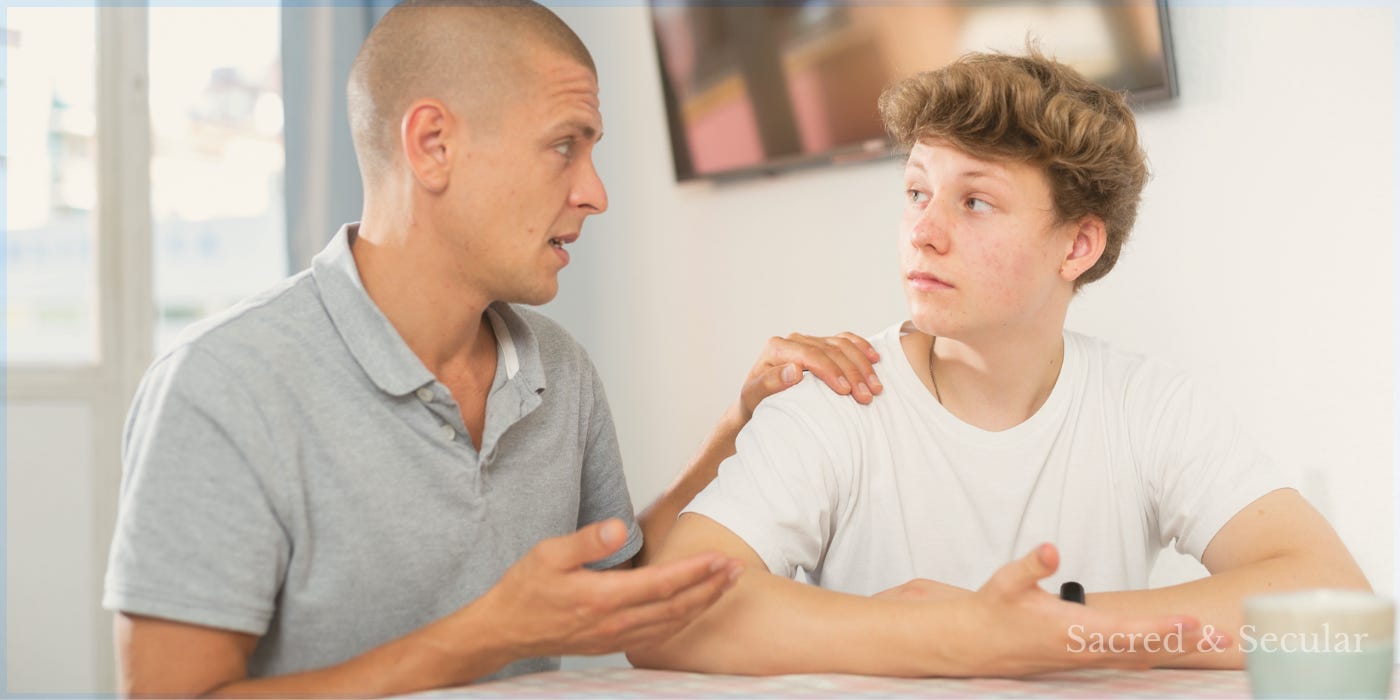 A father and teenage son sitting opposite each other at a kitchen table, talking quietly in warm natural light.