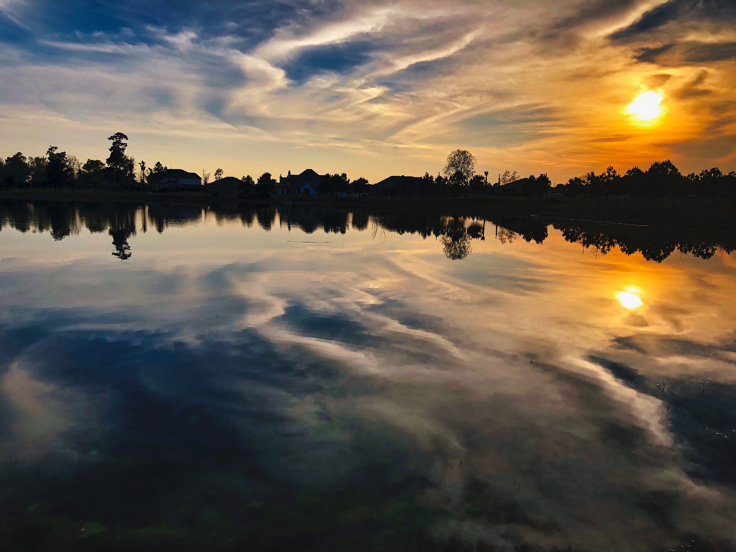 Sunset over the lake with the orangy sun and clouds to the left, the silhouette of trees and houses on the horizon, and the reflection of the clouds and sun in the lake