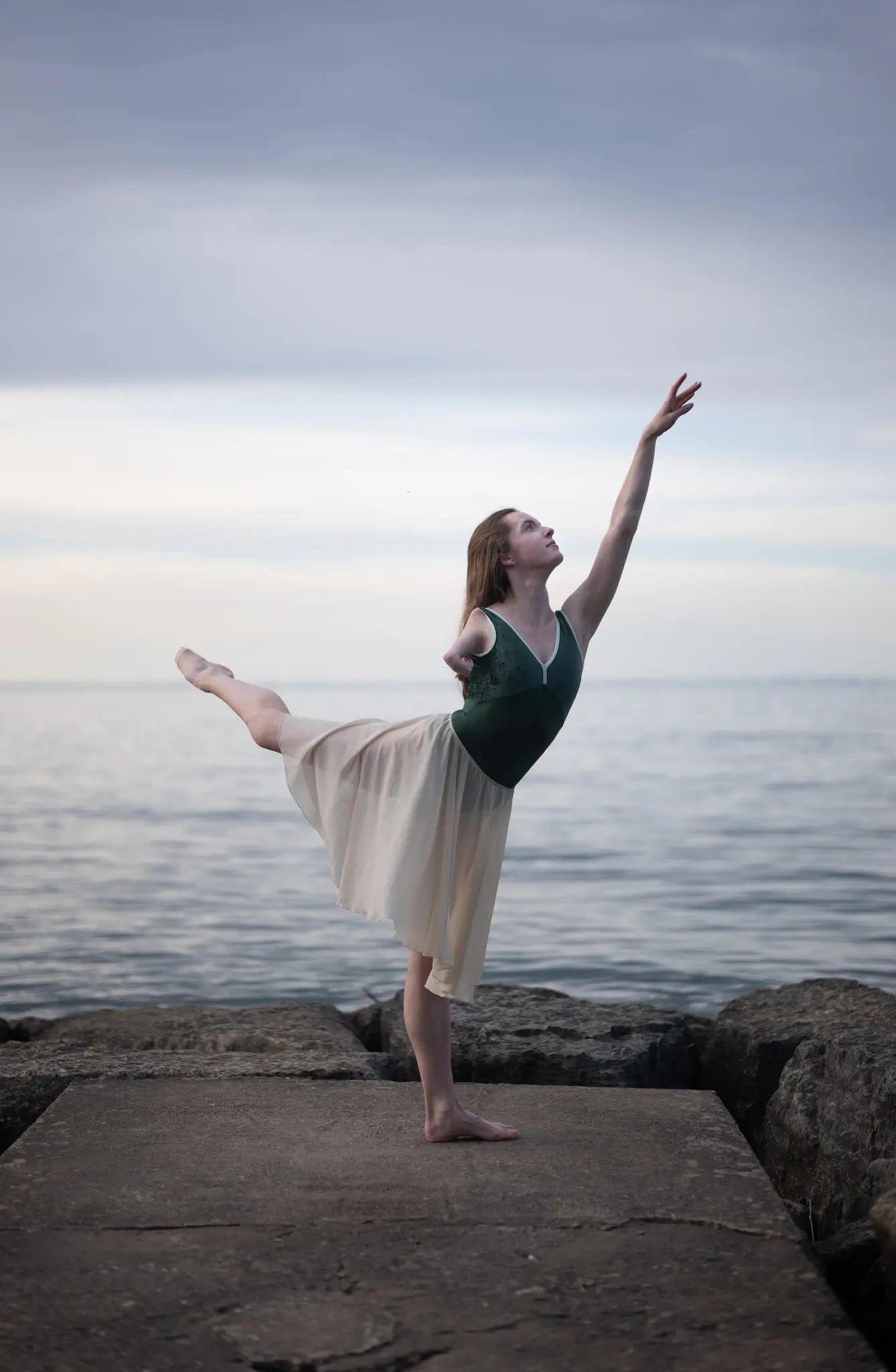 A dancer does an arabesque on a flat surface by a lake. She has one arm extended. Her other arm is short because she has a congenital limb difference.