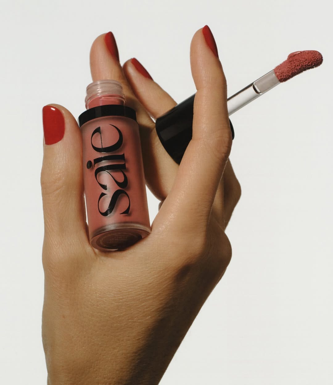 A hand with red nails holding Saie lip gloss.