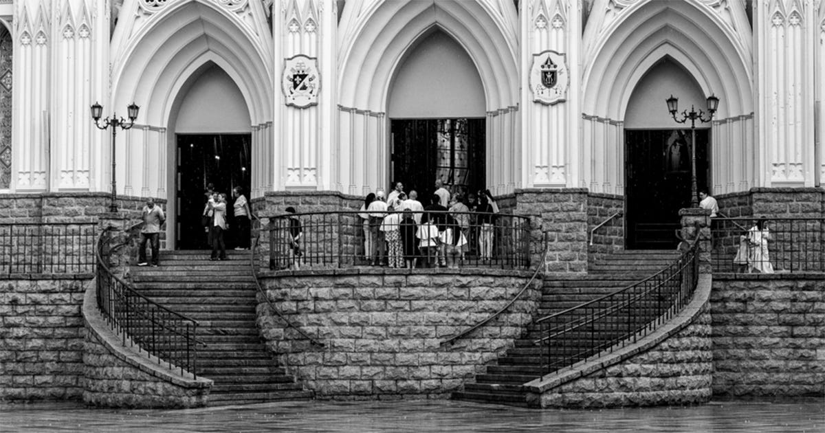A imagem mostra uma paisagem urbana com duas igrejas de estilos arquitetônicos distintos. Em primeiro plano, há uma rua e edificações baixas. Ao fundo, destacam-se os edifícios religiosos, com torres e elementos arquitetônicos visíveis contra o céu. A imagem mostra uma paisagem urbana com duas igrejas de estilos arquitetônicos distintos. Em primeiro plano, há uma rua e edificações baixas. Ao fundo, destacam-se os edifícios religiosos, com torres e elementos arquitetônicos visíveis contra o céu.