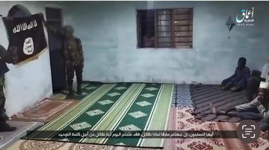 Several figures in dark clothing and head coverings stand and sit on green patterned mats inside a simple room with white walls, barred windows, and Arabic text on the walls. A black flag with white Arabic script is displayed prominently on the wall near the door. The setting appears to be an interior space with a low ceiling and minimal furnishings.