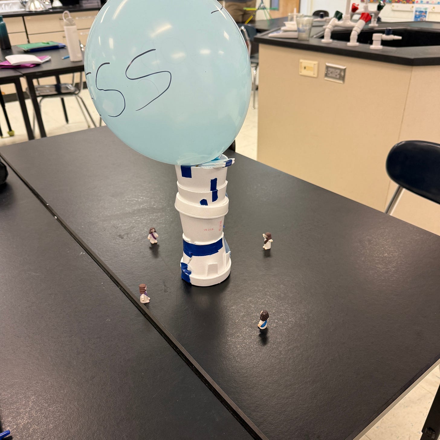 An egg-drop project named God Bless sits on a classroom lab table. The vehicle consists of stacked foam cups and blue tape supporting a large inflated balloon, designed to protect an egg during a fall. Four tiny plastic Jesus figurines stand around the base with their arms outstretched, appearing to bless the vehicle before launch. The model was created by physics students as part of an engineering challenge to safely land an "egg-stronaut" from a height of fifteen feet. An egg-drop project named God Bless sits on a classroom lab table. The vehicle consists of stacked foam cups and blue tape supporting a large inflated balloon, designed to protect an egg during a fall. Four tiny plastic Jesus figurines stand around the base with their arms outstretched, appearing to bless the vehicle before launch. The model was created by physics students as part of an engineering challenge to safely land an "egg-stronaut" from a height of fifteen feet.