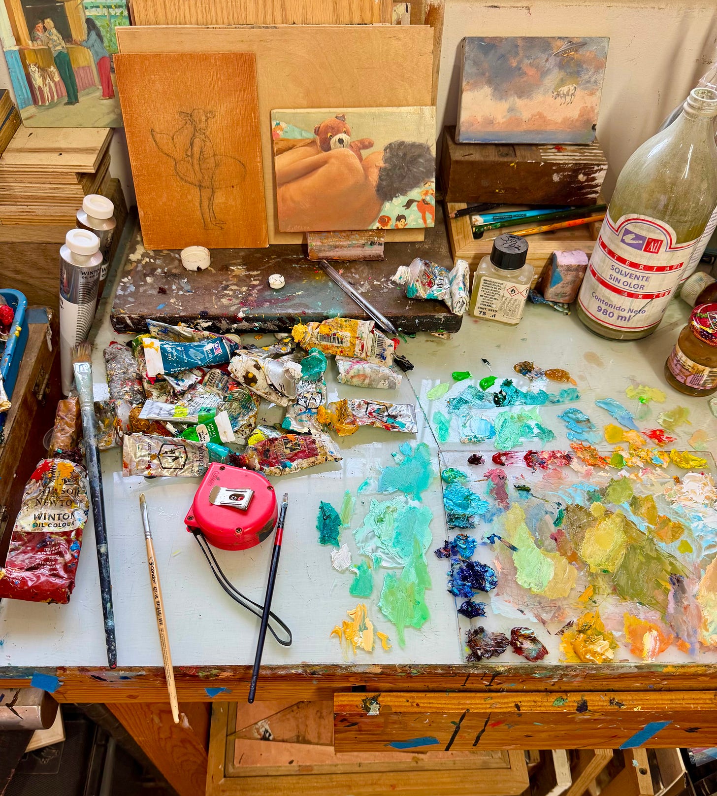 Artist’s worktable covered in paint tubes, brushes, a tape measure, and a palette of mixed greens, blues, yellows, and reds.