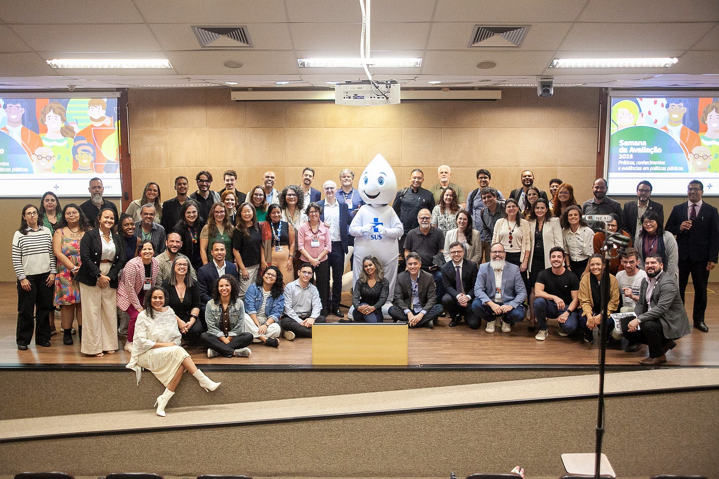Cerca de 50 pessoas, entre homens e mulheres, estão reunidas no palco de um auditório, posando para foto. Algumas estão em pé, enquanto outras estão agachadas ou sentadas. No centro do grupo, está o personagem Zé Gotinha. Ao fundo, dois telões exibem o card do evento.