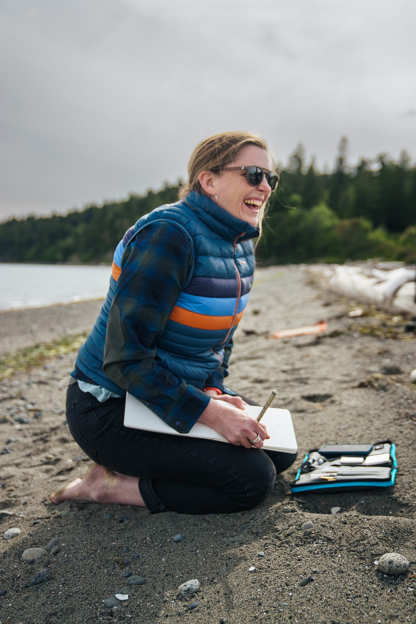 A photo of the artist Anna Brones, sitting on a sandy beach with trees and water behind her, and a notebook and pen in her hand, laughing and smiling 
