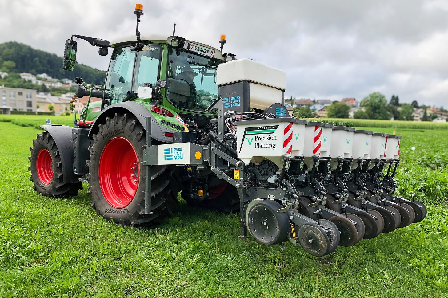 Moderner Fendt-Traktor mit roter Felge und angebauter Einzelkornsämaschine steht auf einem grünen Feld unter grauem Himmel. Am Heck sind mehrere Saatreihenaggregate mit weissen Saatgutbehältern montiert; auf der Maschine stehen die Logos „Swiss Future Farm“ und „Precision Planting“. Moderner Fendt-Traktor mit roter Felge und angebauter Einzelkornsämaschine steht auf einem grünen Feld unter grauem Himmel. Am Heck sind mehrere Saatreihenaggregate mit weissen Saatgutbehältern montiert; auf der Maschine stehen die Logos „Swiss Future Farm“ und „Precision Planting“.