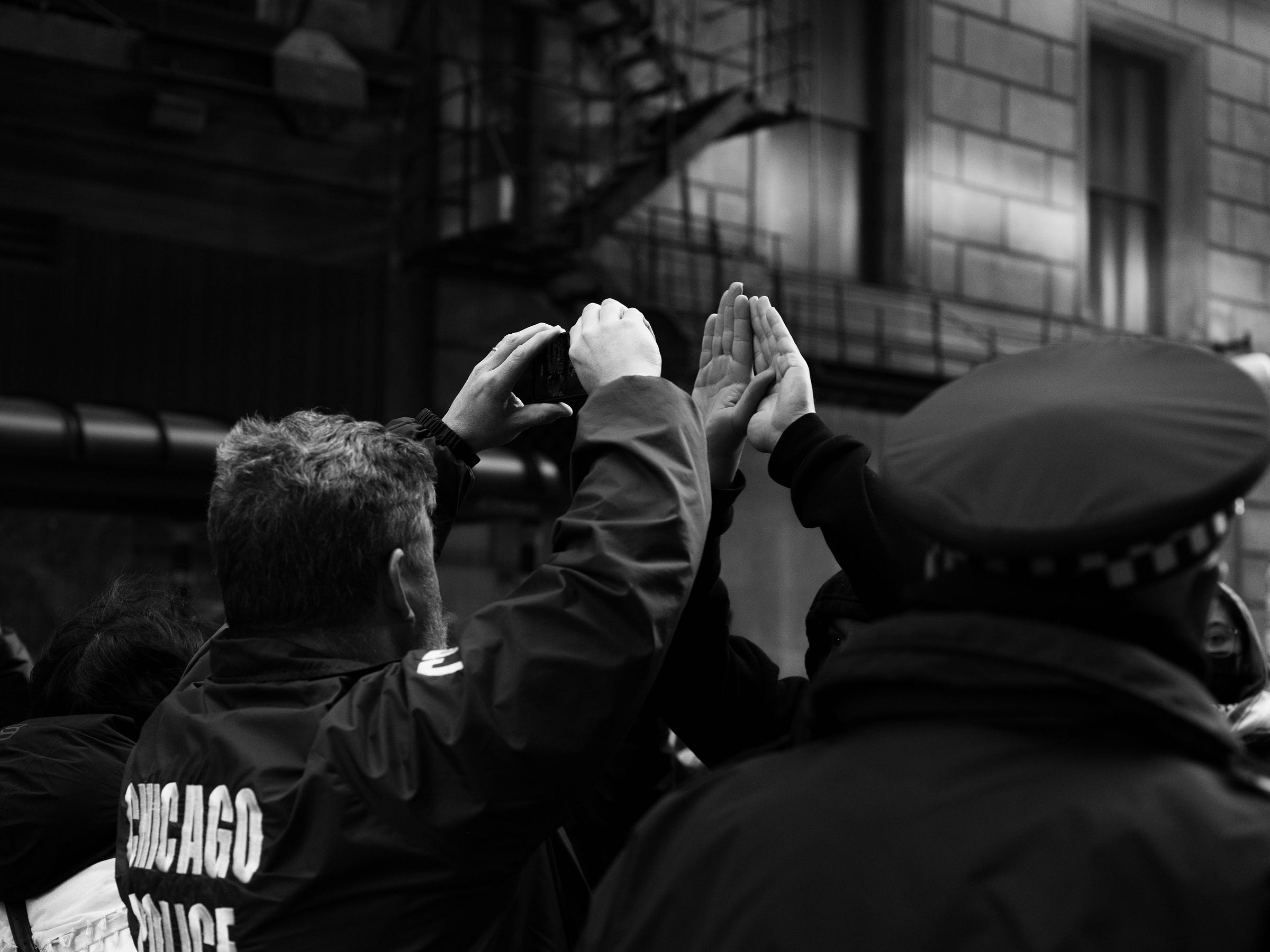 A black-and-white photograph capturing a moment of tension between protesters and Chicago police officers. In the foreground, an officer in a uniform with "Chicago Police" on the back observes the scene, while another officer raises their hands. A protester’s hands are raised in a gesture of defiance or resistance. The blurred urban backdrop, with fire escapes and city architecture, adds to the intensity of the scene, highlighting the standoff between authority and the people.