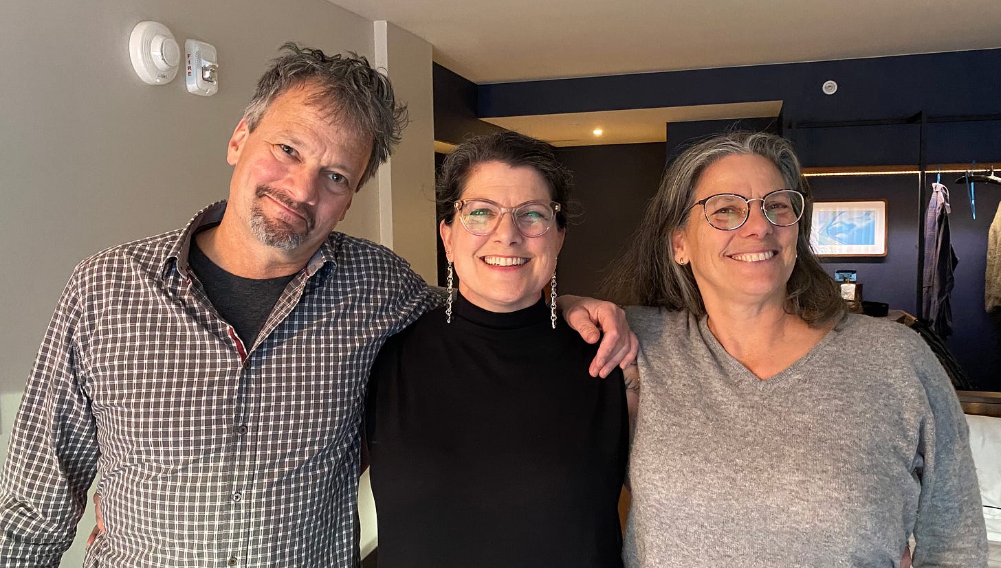 Photo in a hotel room of three siblings: a bearded man in a checked shirt, the author in a black dress, and a gray haired woman in a gray sweater. All smiling. Photo in a hotel room of three siblings: a bearded man in a checked shirt, the author in a black dress, and a gray haired woman in a gray sweater. All smiling.