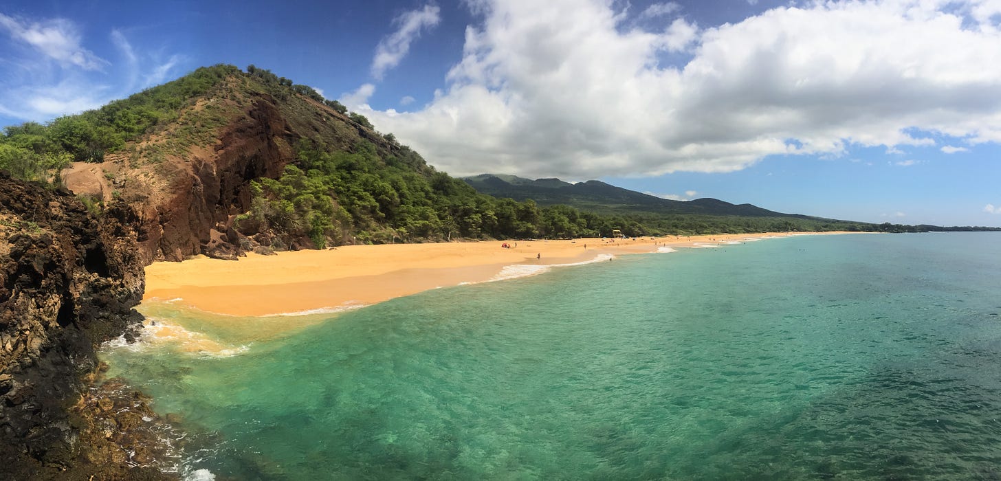 A wide panorama of Big Beach at Mākena State Park, Maui—a long crescent of golden sand meeting clear turquoise water, with a forested cinder cone and lava headland rising on the left under a bright, cloud-scattered sky. A wide panorama of Big Beach at Mākena State Park, Maui—a long crescent of golden sand meeting clear turquoise water, with a forested cinder cone and lava headland rising on the left under a bright, cloud-scattered sky.