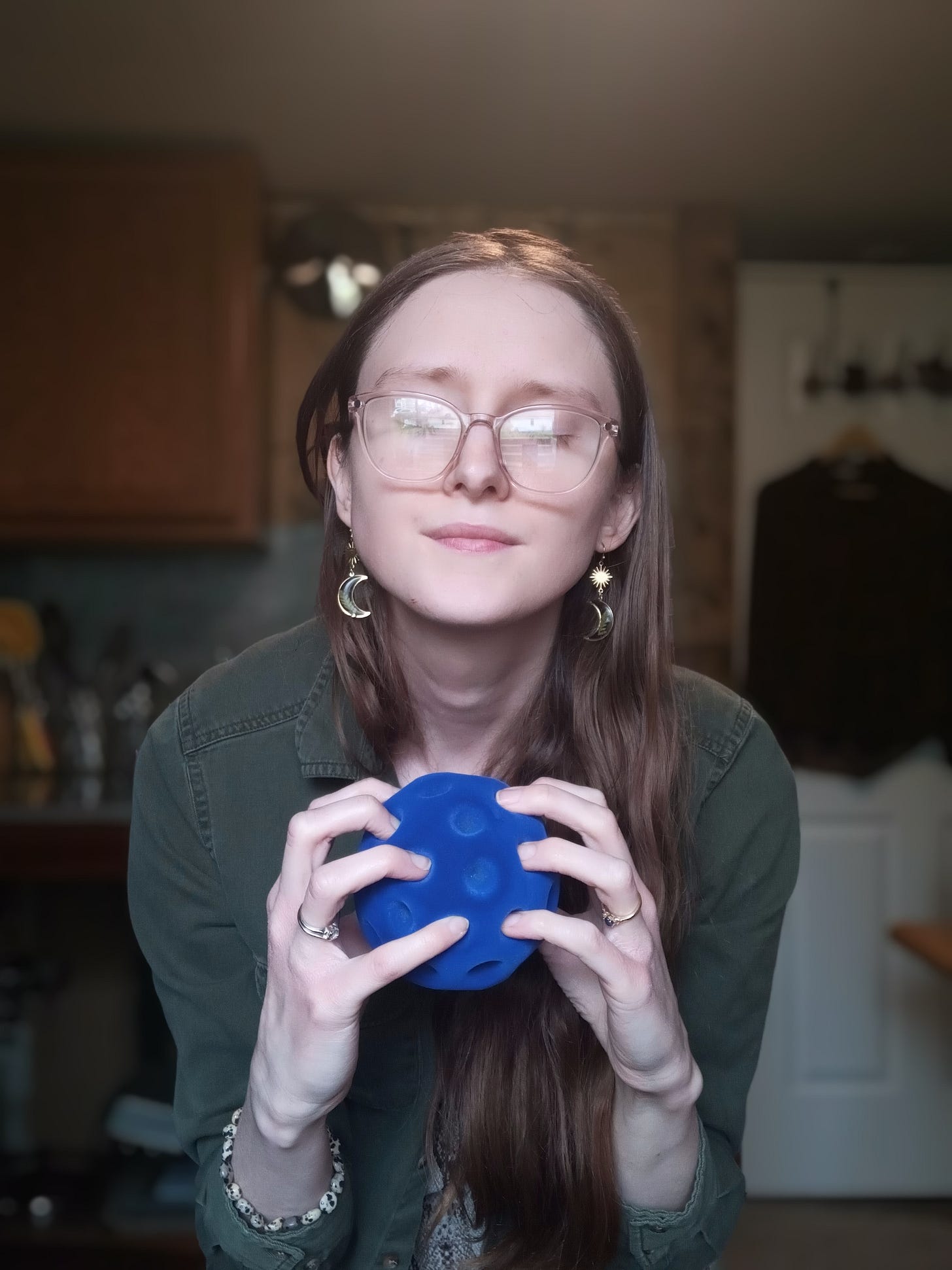 Kandi Zeller (she/her), a white woman with long reddish brown hair and pink glasses, holds a blue Rubbabu stress ball that looks like an oversized golf ball. Kandi has her eyes shut and appears calm. Kandi Zeller (she/her), a white woman with long reddish brown hair and pink glasses, holds a blue Rubbabu stress ball that looks like an oversized golf ball. Kandi has her eyes shut and appears calm.