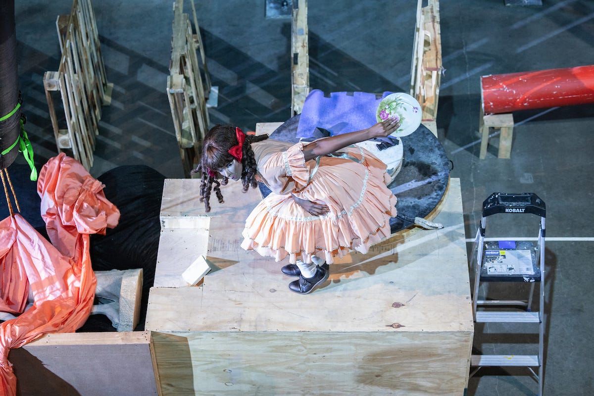 A performer wearing a mannequin mask, light pink dress, and ringlet curls poses on a wooden platform. She crouches forward with one arm resting on her thighs and the other extending straight back, holding a dinner plate. A performer wearing a mannequin mask, light pink dress, and ringlet curls poses on a wooden platform. She crouches forward with one arm resting on her thighs and the other extending straight back, holding a dinner plate.