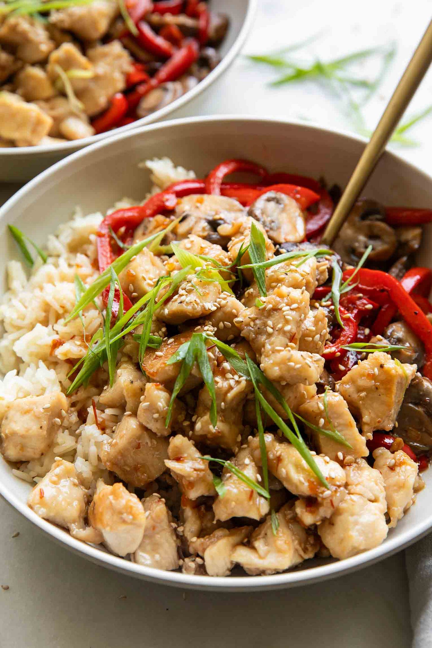 A bowl of rice topped with general tso's chicken, mushrooms, pepper, scallions, and white sesame seeds, a fork sticking out.