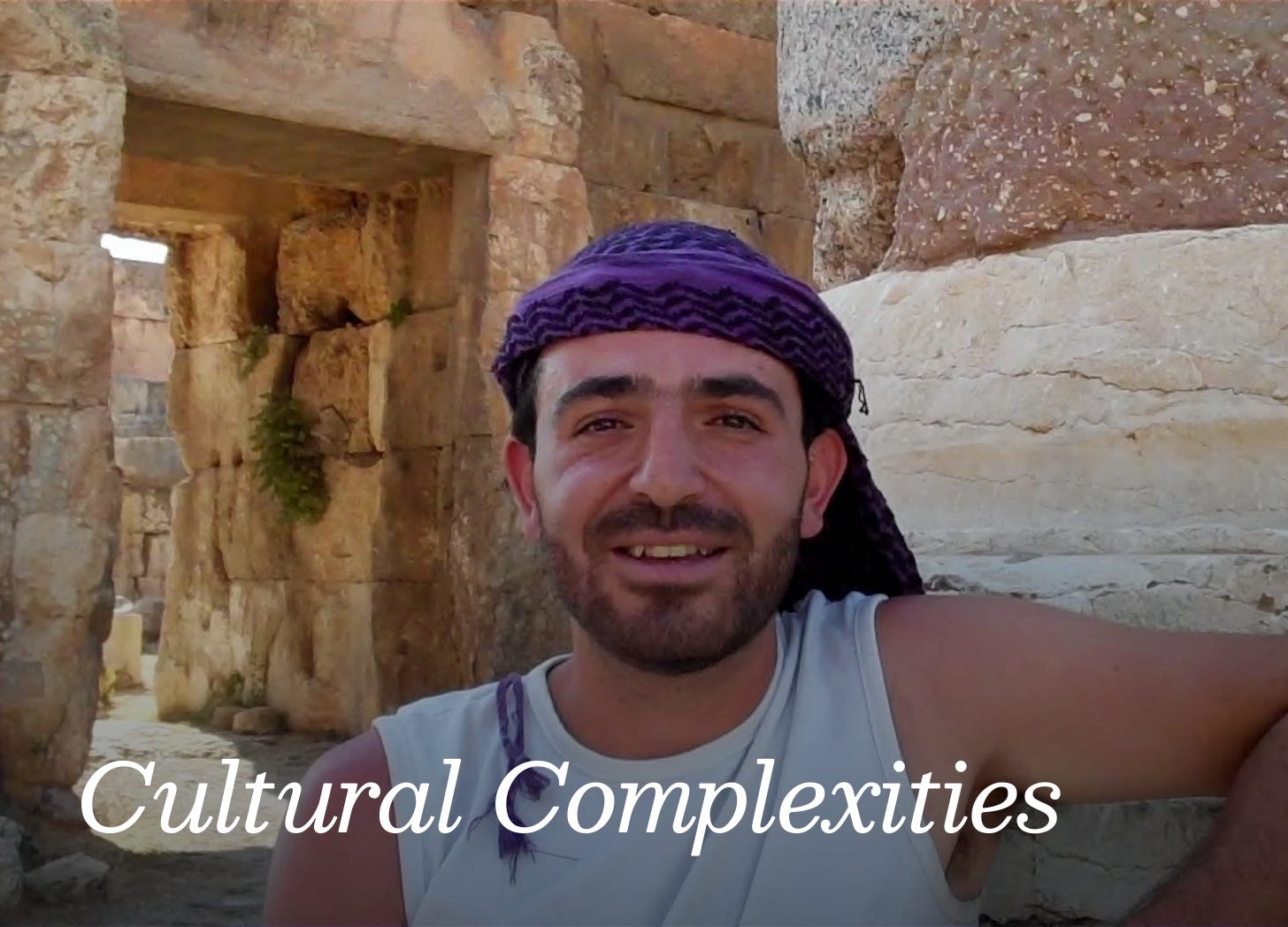 An Arab man wearing a white tank top and purple and black keffyeh. He is outside amid ancient stone buildings, standing casually and smiling slightly. 