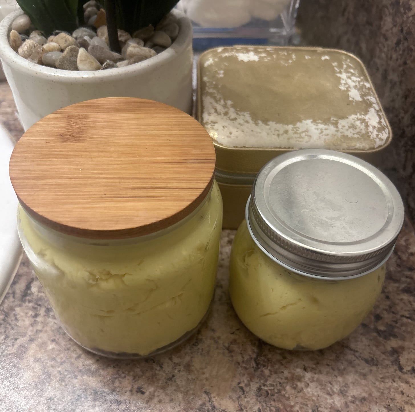 Two glass jars filled with homemade, whipped yellow shea body butter on a speckled countertop. One jar features a bamboo lid and the other a silver metal lid, showcasing a rich, thick, natural skincare texture.