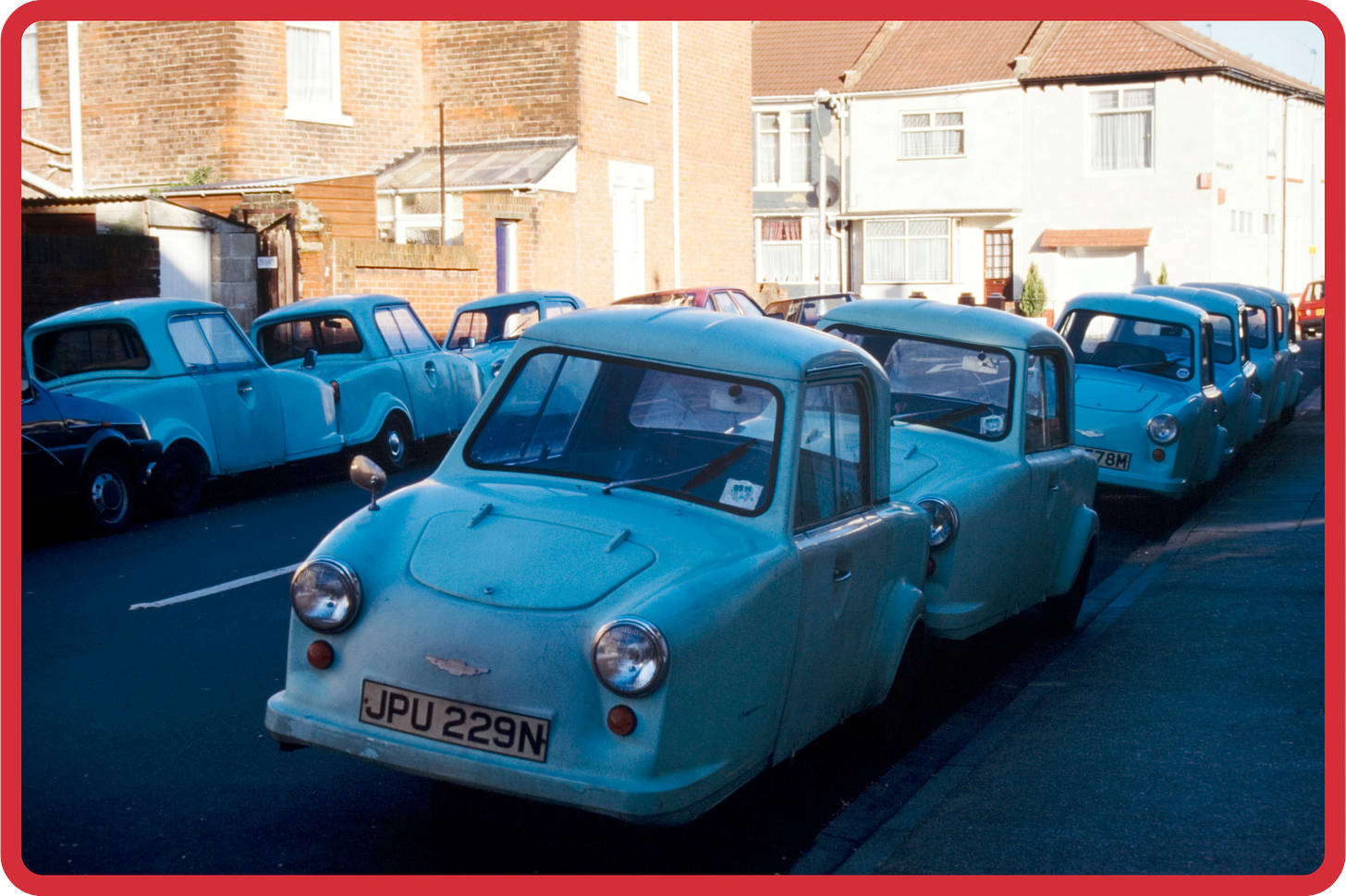 A vintage photo of around a dozen blue invalid carriages parked on a residential street. The carriages are mostly facing towards us, showing their distinctive long bonnets. In the background, a few suburban townhouses are lit by bright sunshine. A vintage photo of around a dozen blue invalid carriages parked on a residential street. The carriages are mostly facing towards us, showing their distinctive long bonnets. In the background, a few suburban townhouses are lit by bright sunshine.