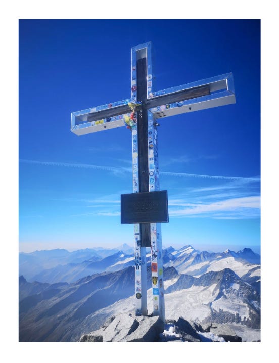 Summit cross on snowy mountain peak with mountain range backdrop.