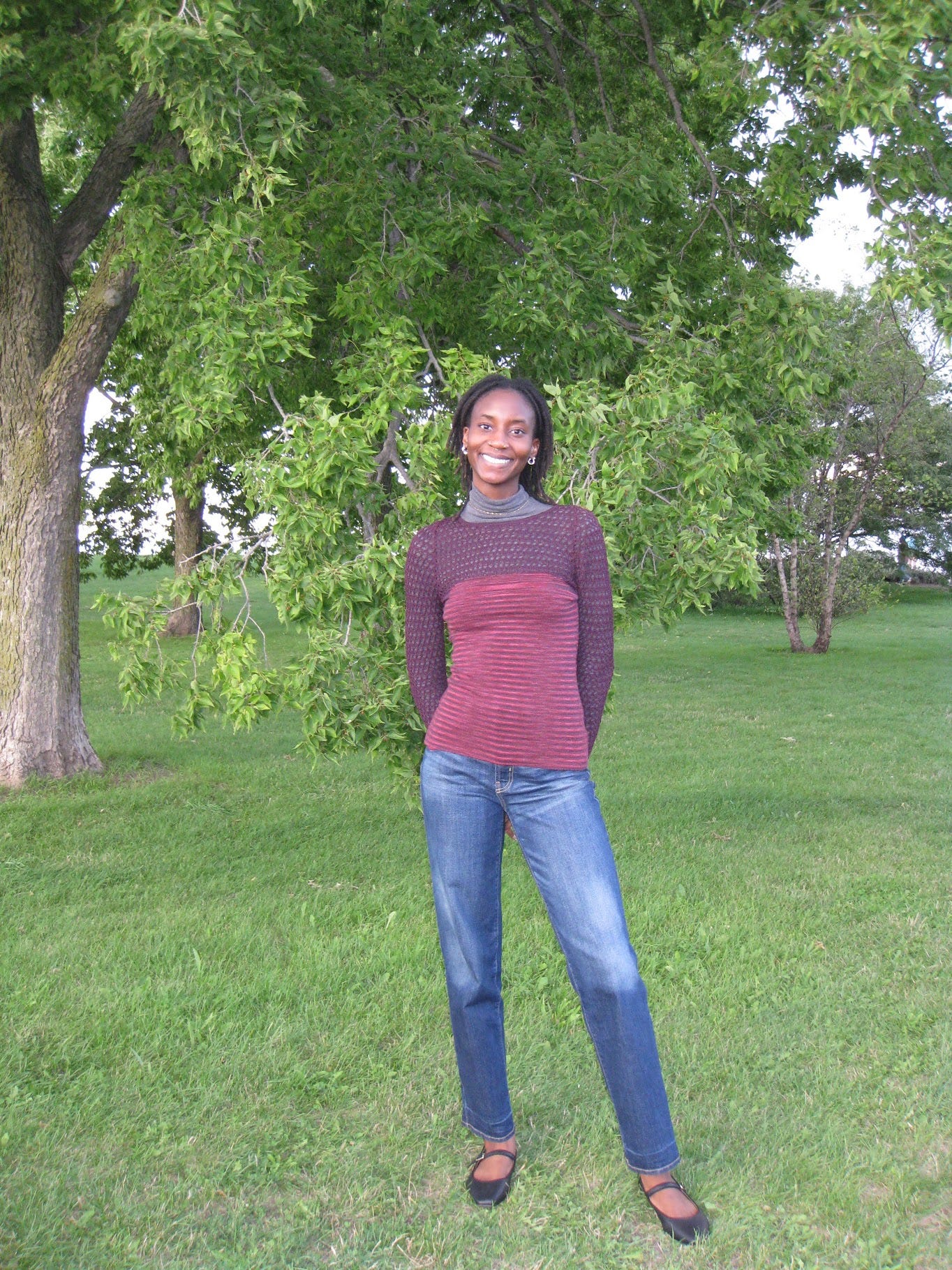 A photo of me standing before a tree in the same stripy, mesh outfit with my jeans and ballet flats. My hands are behind my back and I am contrapposto..