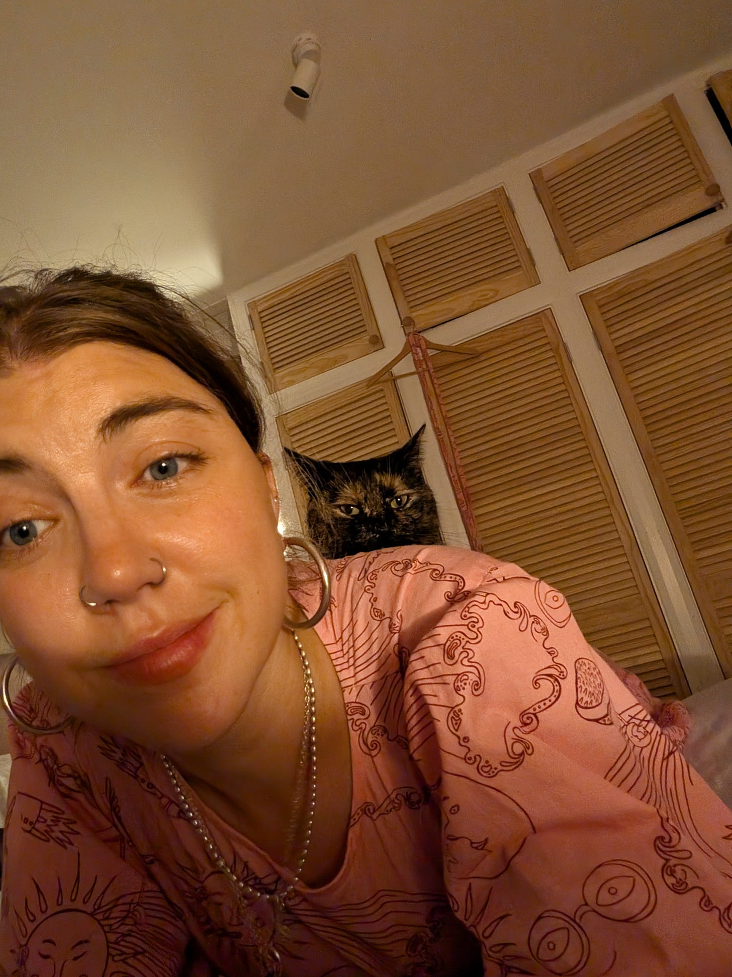 Grace is close-up lying on her bed with wardrobe doors visible behind her; the cat is peering over her shoulder with daggers in her eyes. Grace is wearing an off-pink dress with doodle patterns on it, plus silver hoops in her ears and nose.