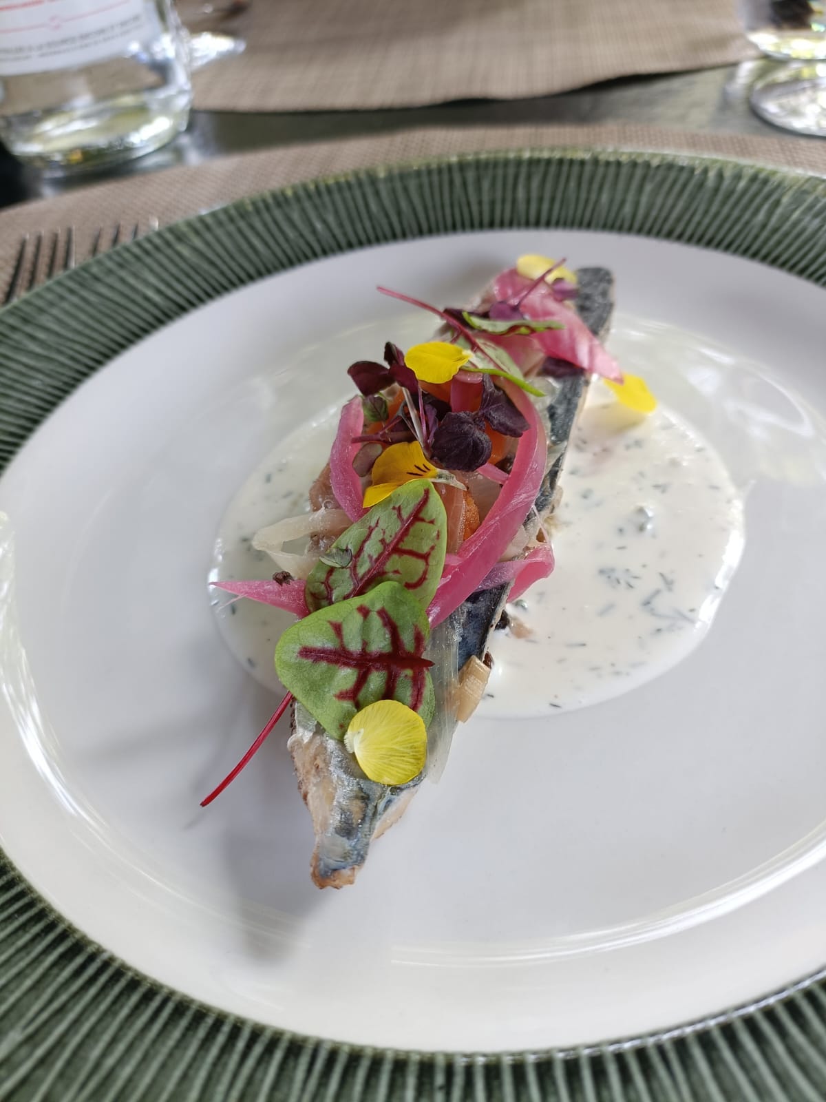A single fillet of fish on a large white plate, topped with pickled red onion, edible flowers and micro greens, in a pool of herb-flecked cream sauce. A single fillet of fish on a large white plate, topped with pickled red onion, edible flowers and micro greens, in a pool of herb-flecked cream sauce.