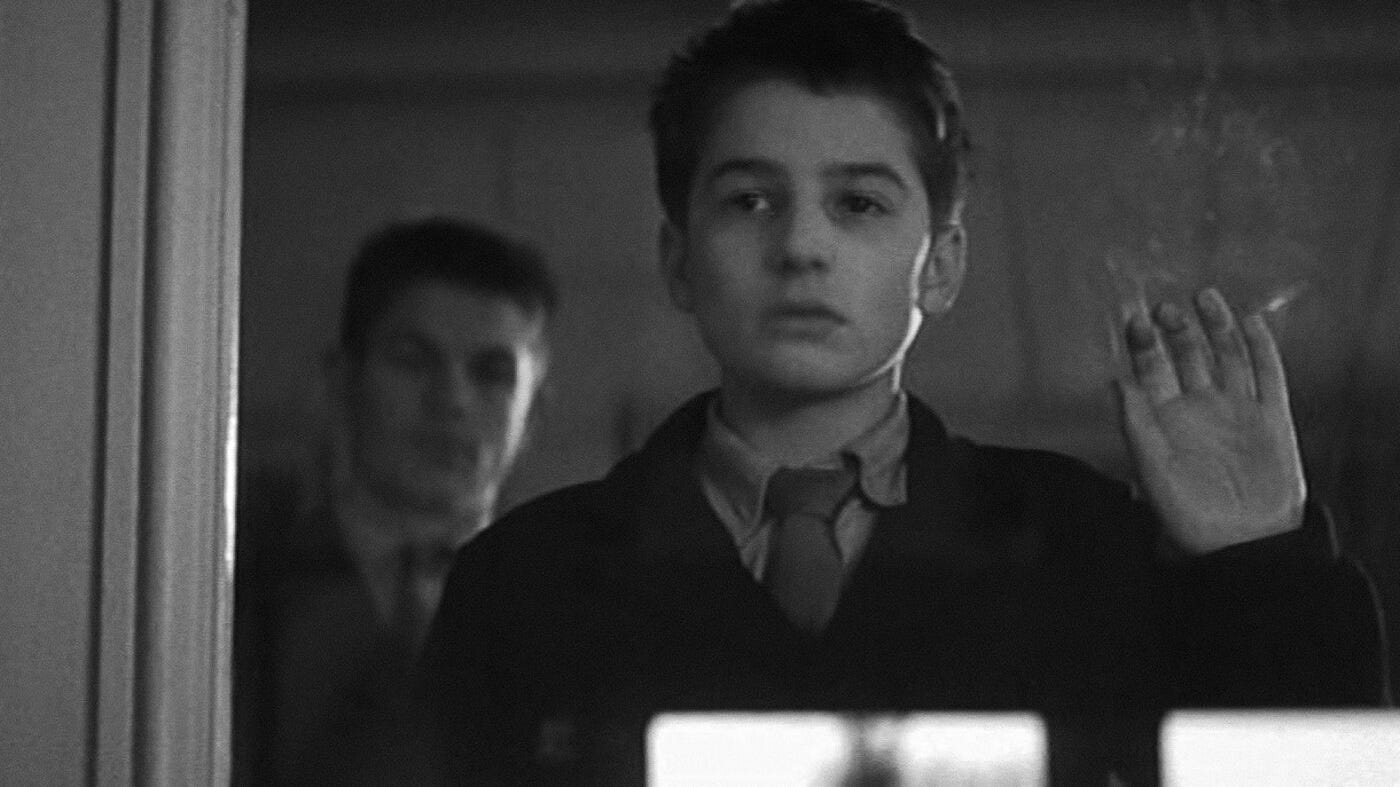 Antoine stands behind a glass window, his hand pressed against it, gazing out with a solemn expression.